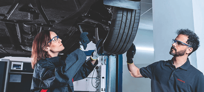 Zwei Nissan-Mechaniker mit Schutzbrillen prüfen einen Reifen an einem angehobenen Fahrzeug in einer Werkstatt.